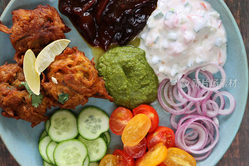 餐盘的全帧图像包含自制的脆洋葱bhajis / pakoras，黄瓜片，樱桃番茄，红洋葱圈，raita和酸辣酱蘸汁，印度菜素食小吃，木纹背景，高架视野